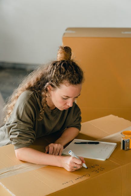A young woman with long brown hair, wearing a grey t-shirt and white pants, is sitting on a light wooden floor inside a room during a home relocation process. She is holding a set of keys in her right