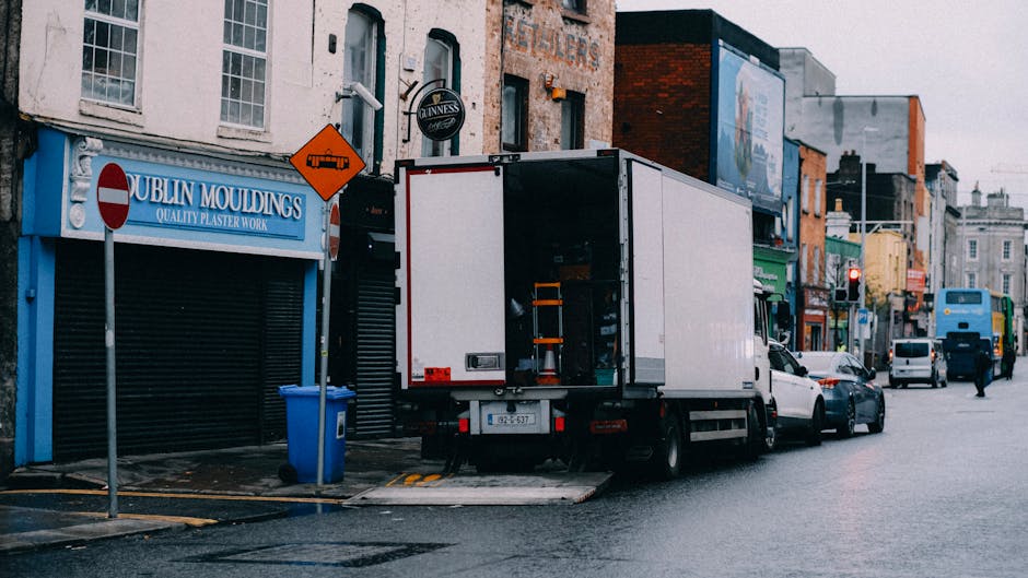 A large white moving truck is parked on a city street outside a commercial building with a sign that reads 'Dublin Mouldings Quality Plaster Work.' The rear door of the truck is open, revealing an int